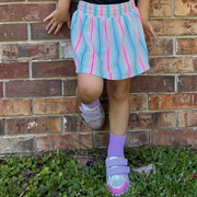 Model stands, leaning up against a brick wall. She has on a bright pink, blue, purple, & peach pinstripe skirt on with purple socks with a pair of Rake multi metallic sneakers. The back sole of the right foot is propped up on the wall.