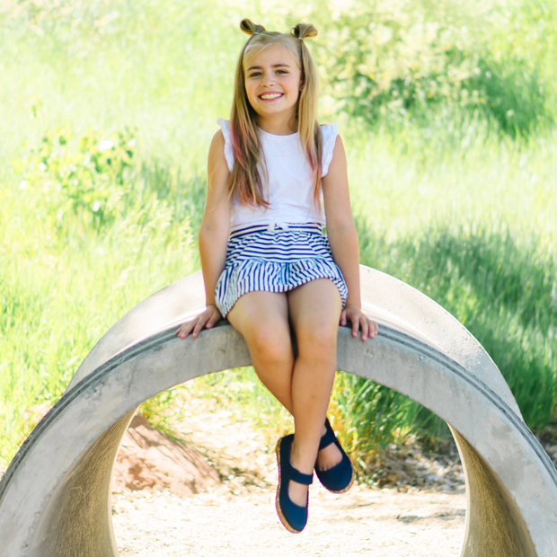 Young white girl with blond hair in bun-pigtails sits on a concrete arch outside on a sunny day. She is wearing a white ruffle top, white & navy stripped skirt, and a pair of navy Ravine mary jane seakers