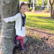 Little white girl with blond hair in a pink bow stands next to a tree hugging it behind her outside in a park. She is wearing a white cable knit caridgan & skirt set with navy details, a navy floral top, long, bright fushcia socks, and a pair of navy Ravine mary jane sneakers