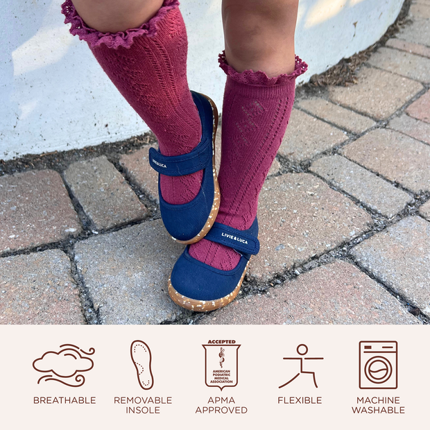 Child seen from the knees down wearing tall, fuscia knitted socks with navy Ravine sneaker mary janes stands outside on paved bricks. Below are icons highlighting the following shoe features; beathable, removeable insole, APMA approved, flexible, machine washable